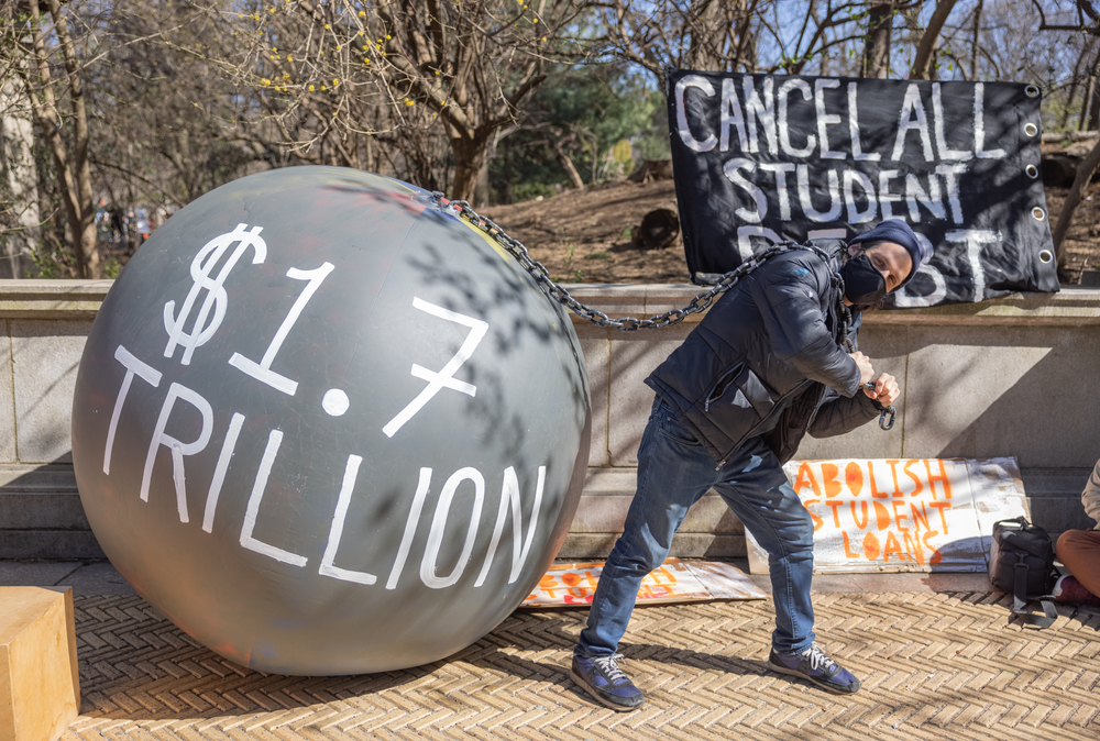 A protester calling for student loan debt relief carries a cartoonish $1.7 Trillion ball and chain.