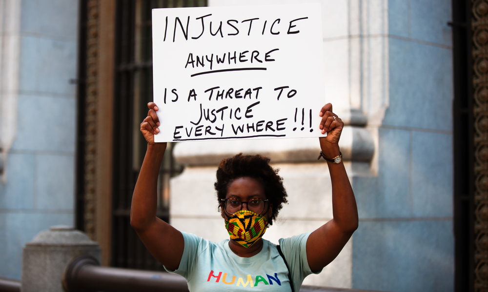 A Black woman holding a protest sign that reads "Injustice anywhere is a threat to justice everywhere!"